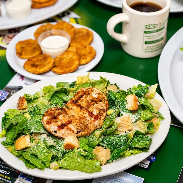 Grilled chicken Caesar salad with croutons and parmesan, alongside fried pickles and coffee on a green table.