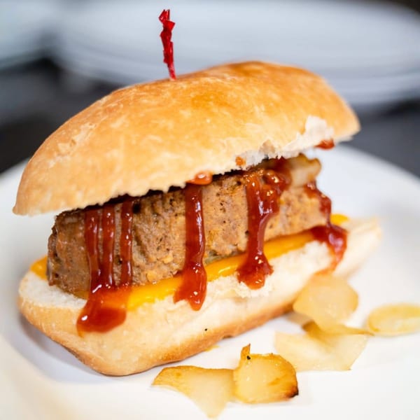 Meatloaf sandwich on a white plate, topped with barbecue sauce and a red toothpick.