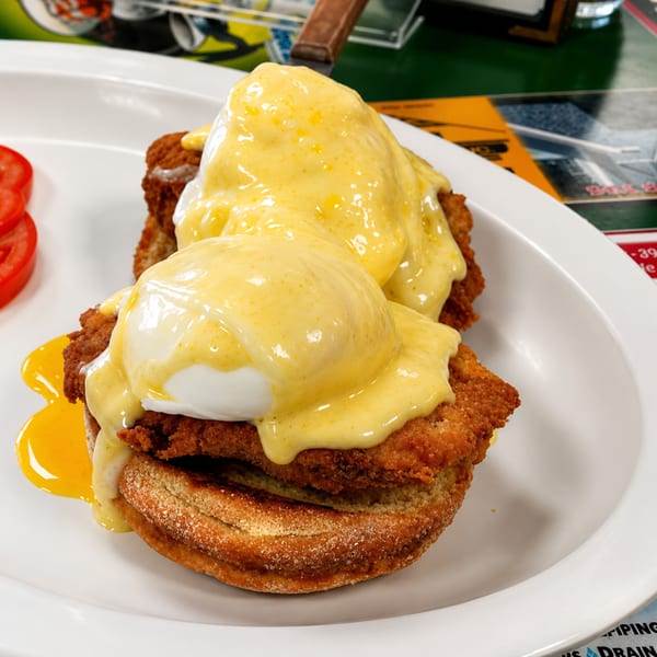 Two fried chicken and poached egg Benedicts with hollandaise sauce and sliced tomatoes on a white plate.