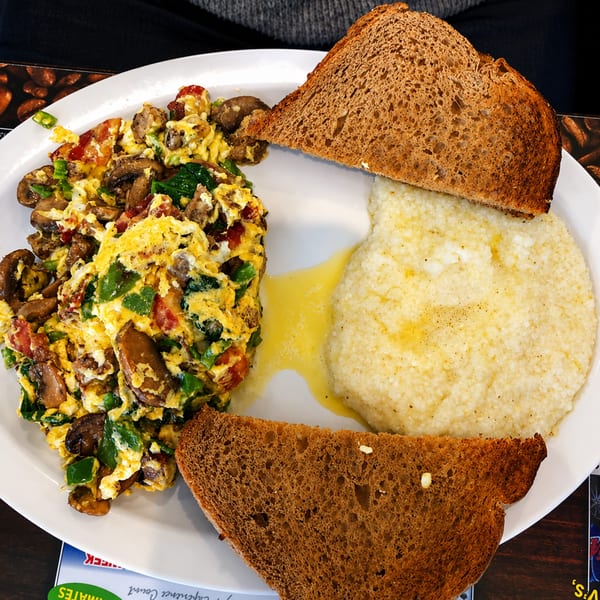 Scrambled eggs with mushrooms, peppers, and tomatoes, served with grits and two slices of toast.