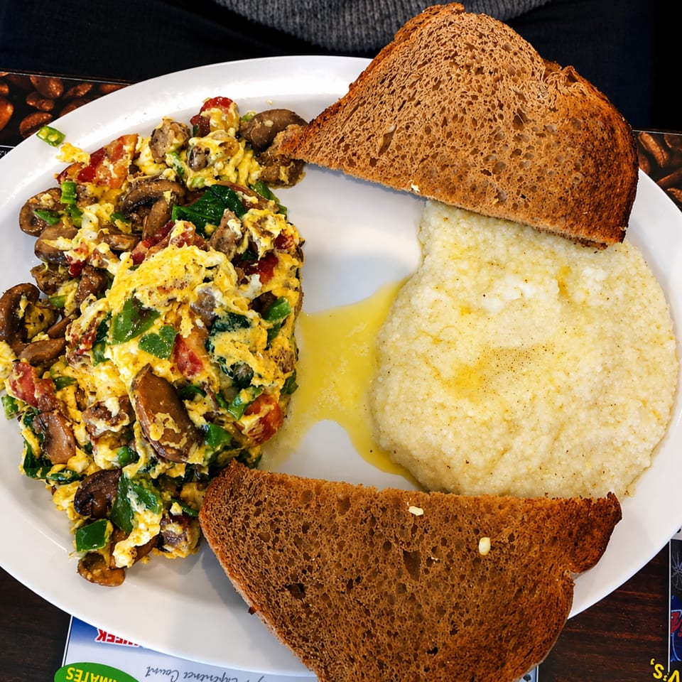 Scrambled eggs with mushrooms, peppers, and tomatoes, served with grits and two slices of toast.